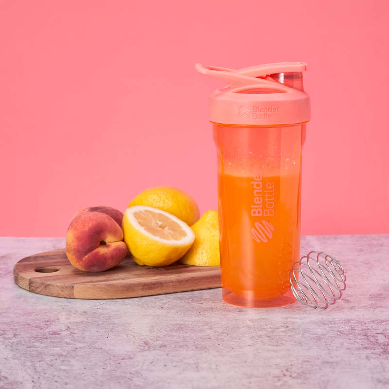 A bottle of an orange protein shake surrounded by fresh fruits, including lemons and a peach, highlighting the citrus ingredients of the drink.