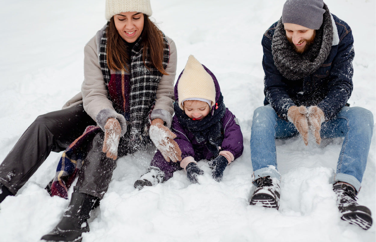 A family of two adults and a child bundled in winter clothing sit in the snow, capturing a joyful winter scene.