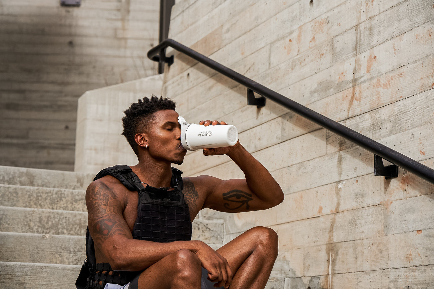 A man in a black vest drinks from a white shaker, eyes closed, with a visible tattooed arm, against a concrete stairwell.