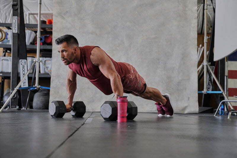 A man performing push-ups using dumbbells indoors, emphasizing strength training and physical fitness with visible exercise equipment and athletic clothing.