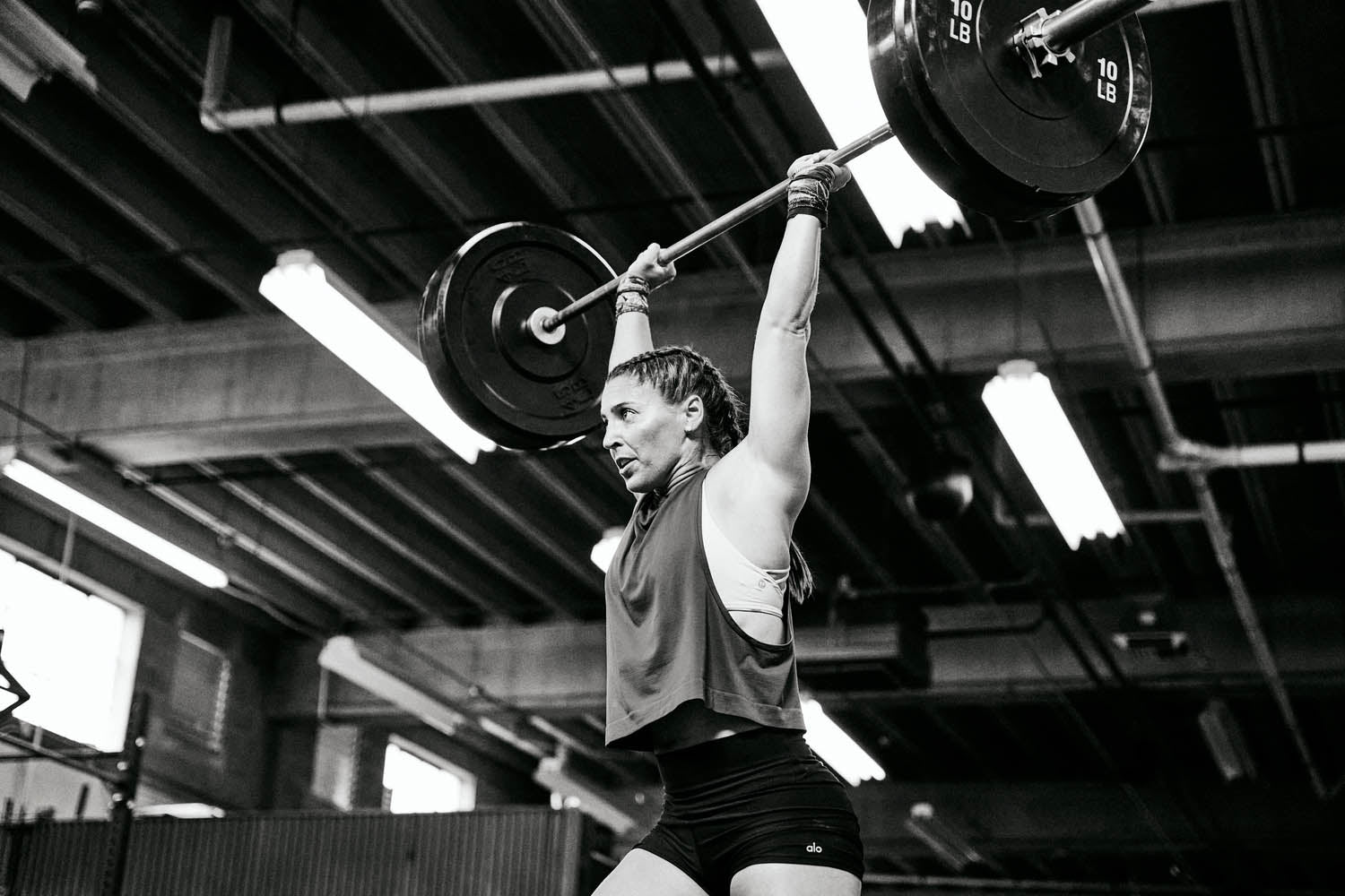 A woman in a gym lifting a barbell, wearing a tank top, focusing on strength training with visible weight plates and fitness equipment.
