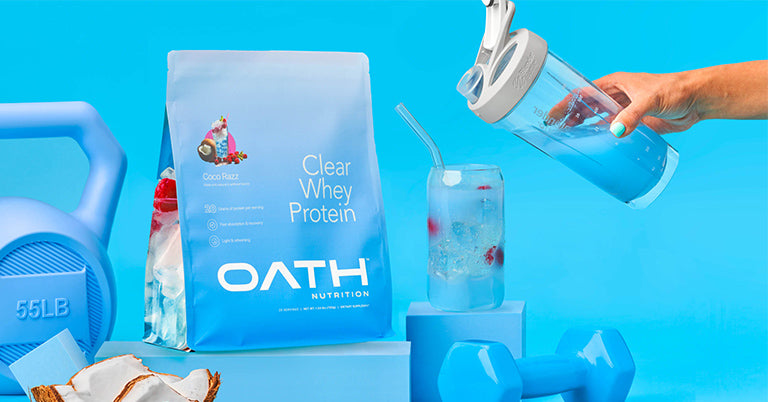 A protein shaker is held by a hand, pouring clear whey protein into a glass cup, with a blue kettlebell partially visible in the background.