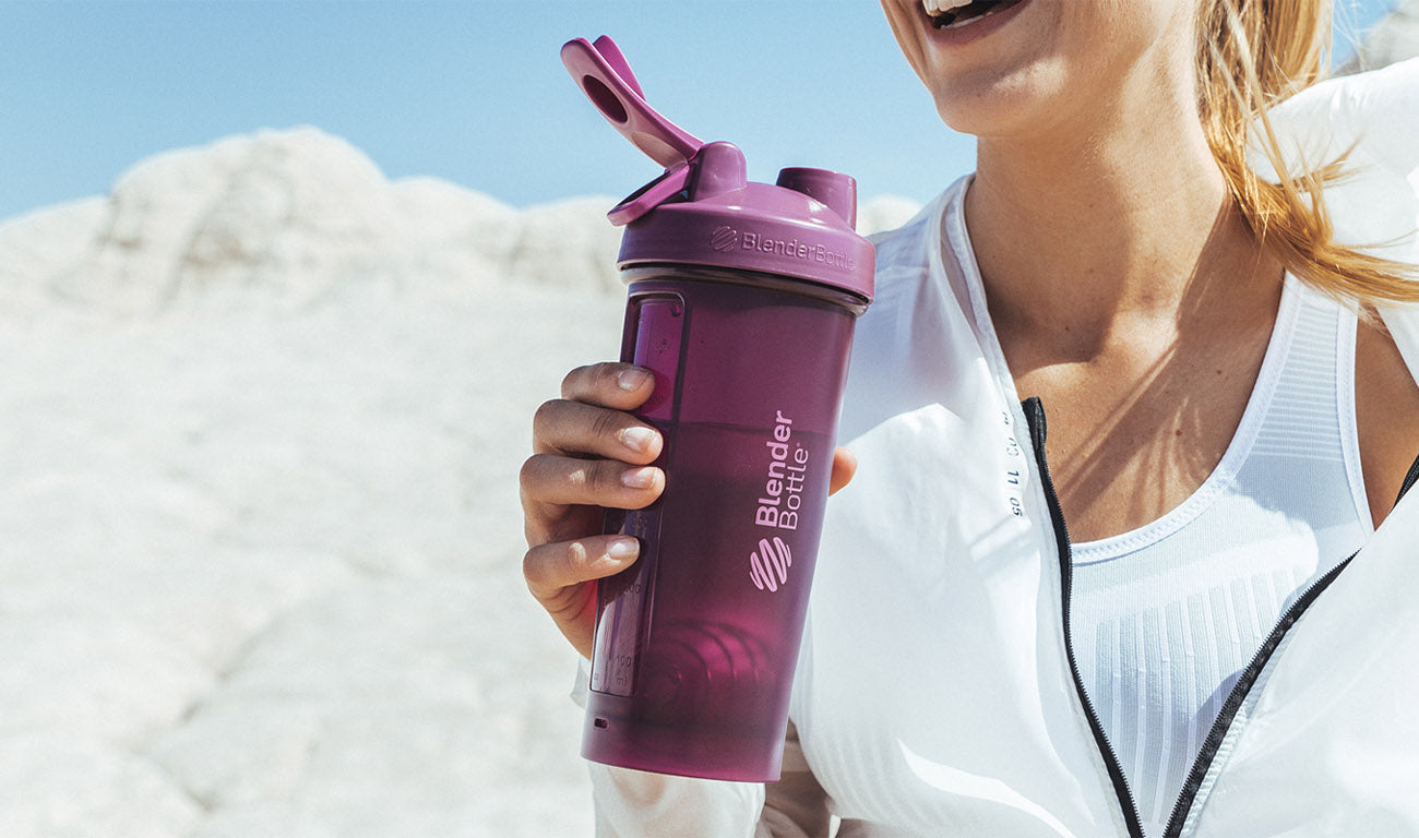 A woman in a white shirt holds a purple BlenderBottle outdoors, suggesting a casual, sunny setting.