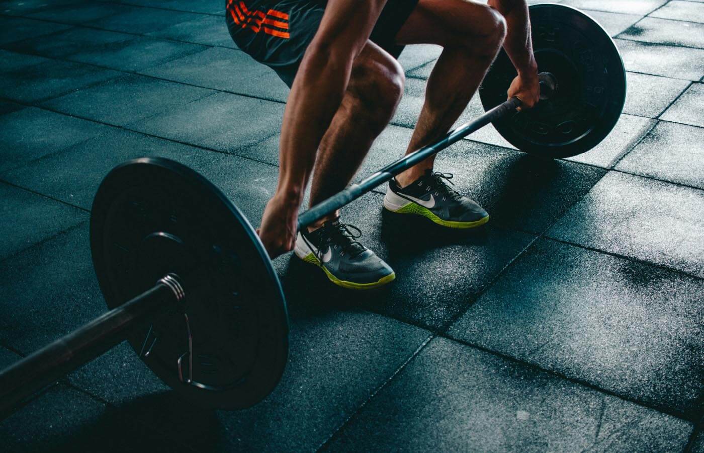 A man lifts weights on a gym floor, showcasing strength training with visible barbell and exercise equipment, wearing athletic footwear.