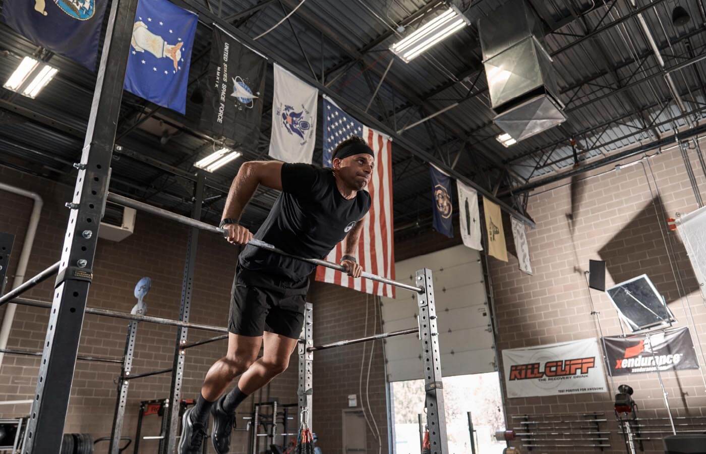A man in a gym doing a pull-up, wearing black shorts and shoes, with a white and blue flag visible in the background.