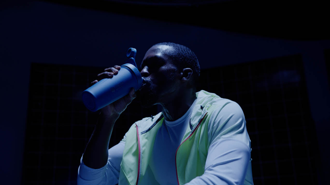 A man in a white shirt and yellow jacket drinks from a blue BlenderBottle shaker.