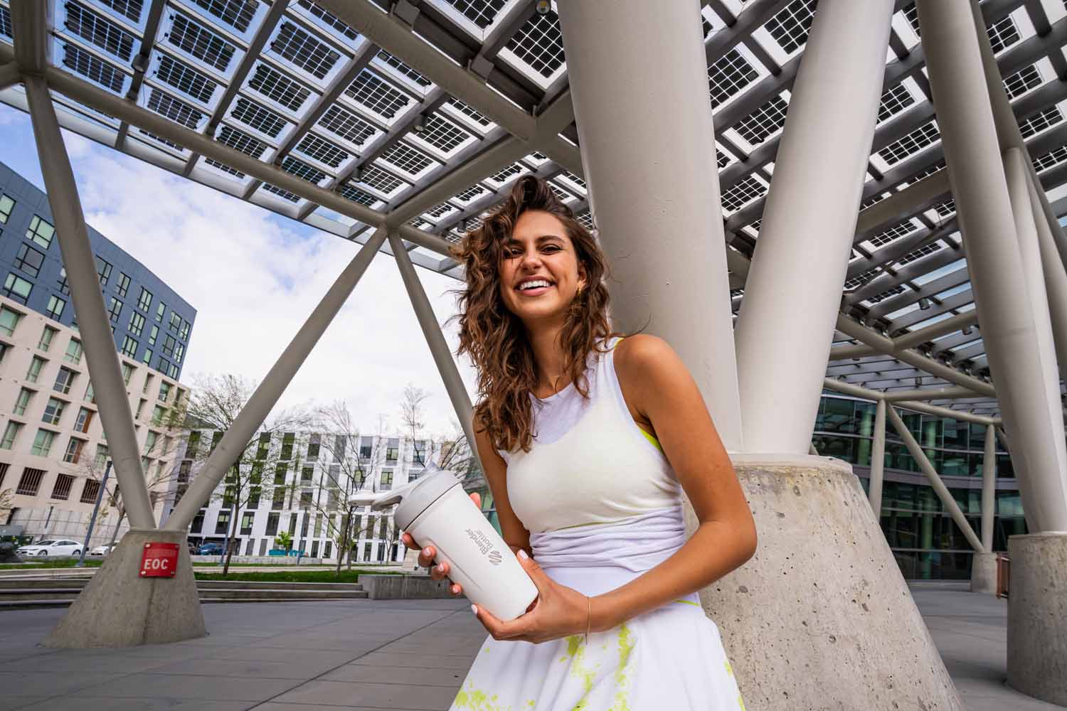 A woman smiles while holding a white BlenderBottle, standing outdoors with a building in the background.