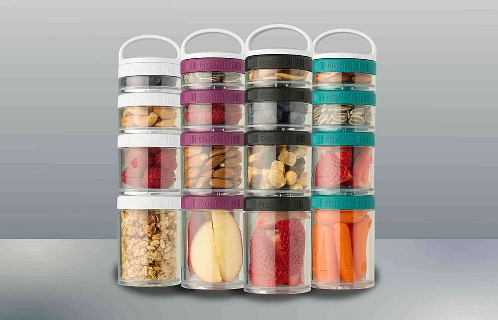 Assorted GoStak jars filled with raspberries, strawberries, nuts, and carrots, alongside a cut apple and cereal, displayed in a neat arrangement.