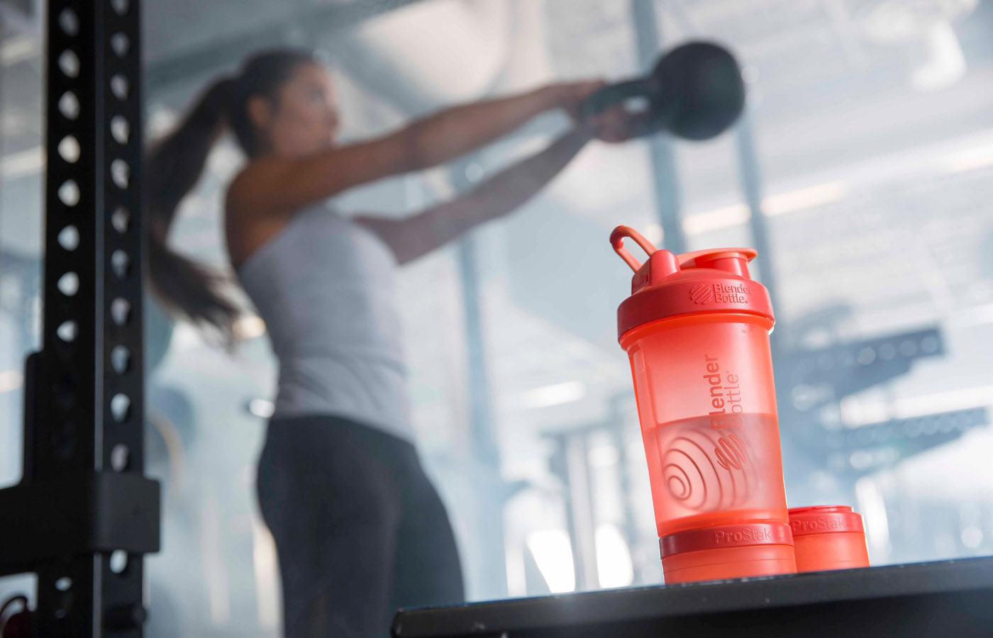 Orange Prostak shaker bottle with a woman lifting a kettlebell in a gym in the background.