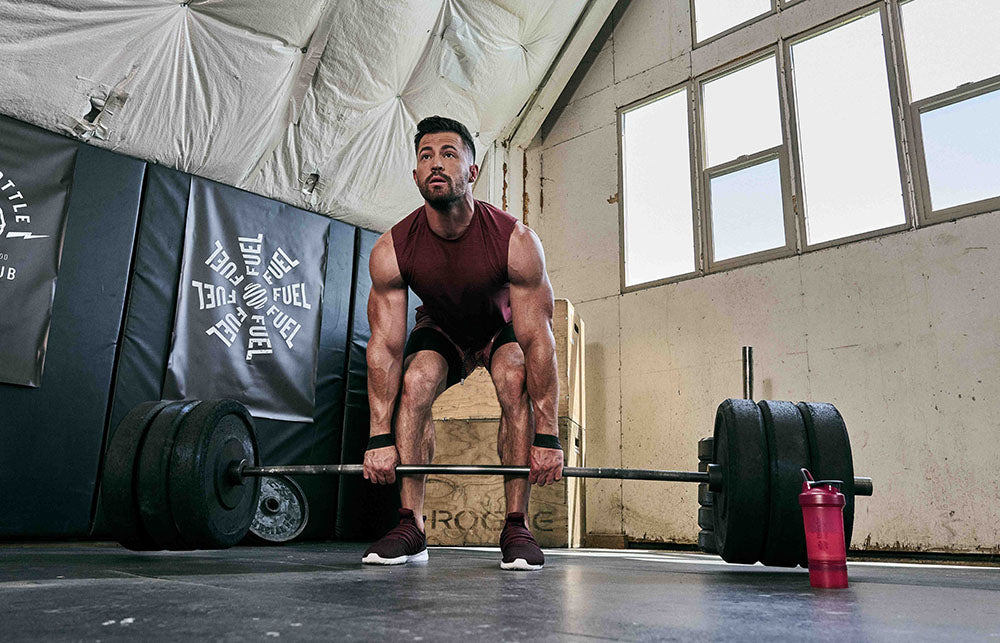 A man in a red tank top lifts weights in a gym, with a red BlenderBottle and exercise equipment visible nearby.