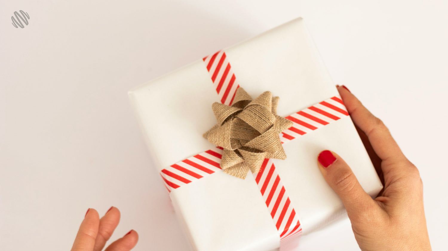 A person holds a gift wrapped with burlap bow and red-striped tape, showcasing fingers and a grey BlenderBottle logo on the packaging.