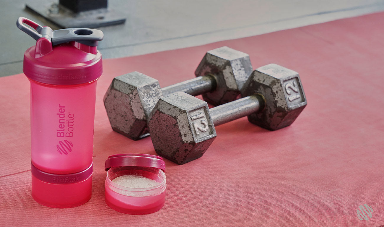 A pair of dumbbells and a pink ProStakshaker bottle on a red surface.