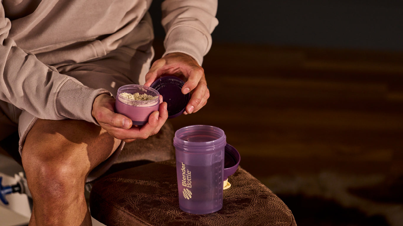 A person holding a close-up of a blender bottle and a container of powder, with a purple plastic bottle visible in the background.