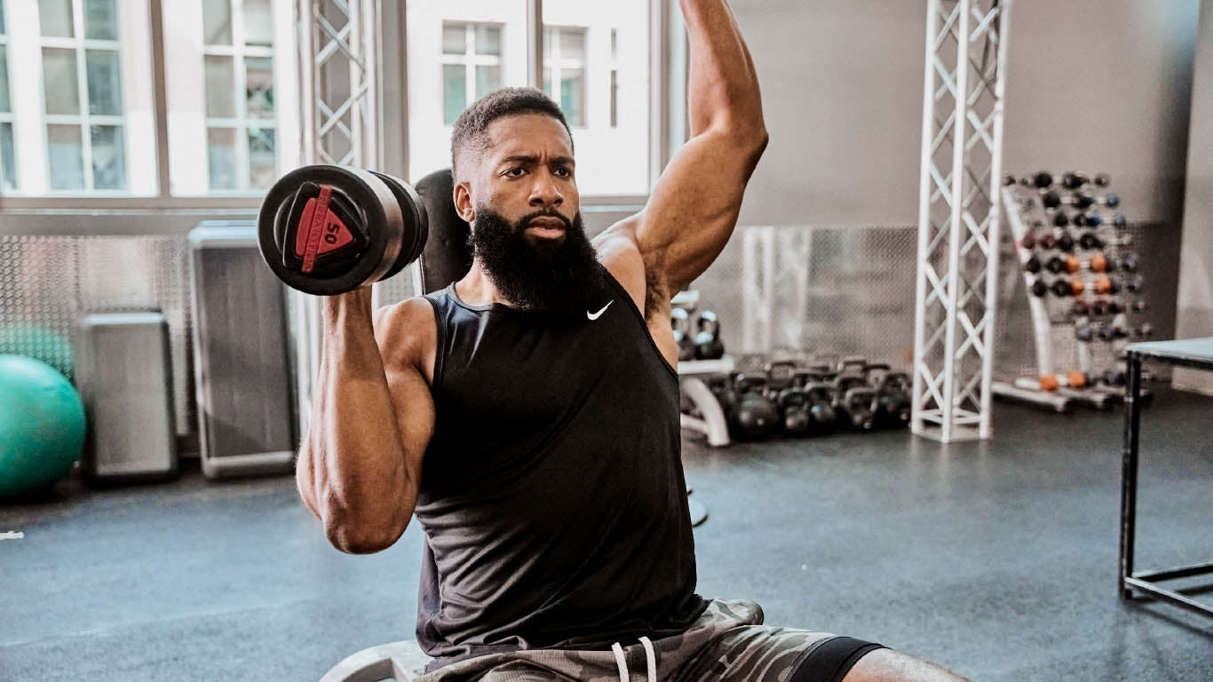 A man lifts weights in a gym, focusing on strength training with exercise equipment visible in the background.