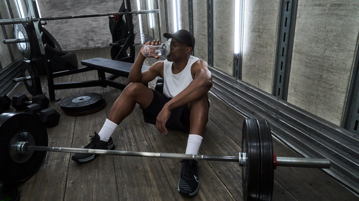 A man sits on a bench drinking water from a BlenderBottle shaker with a black weight plate nearby, in a gym setting.