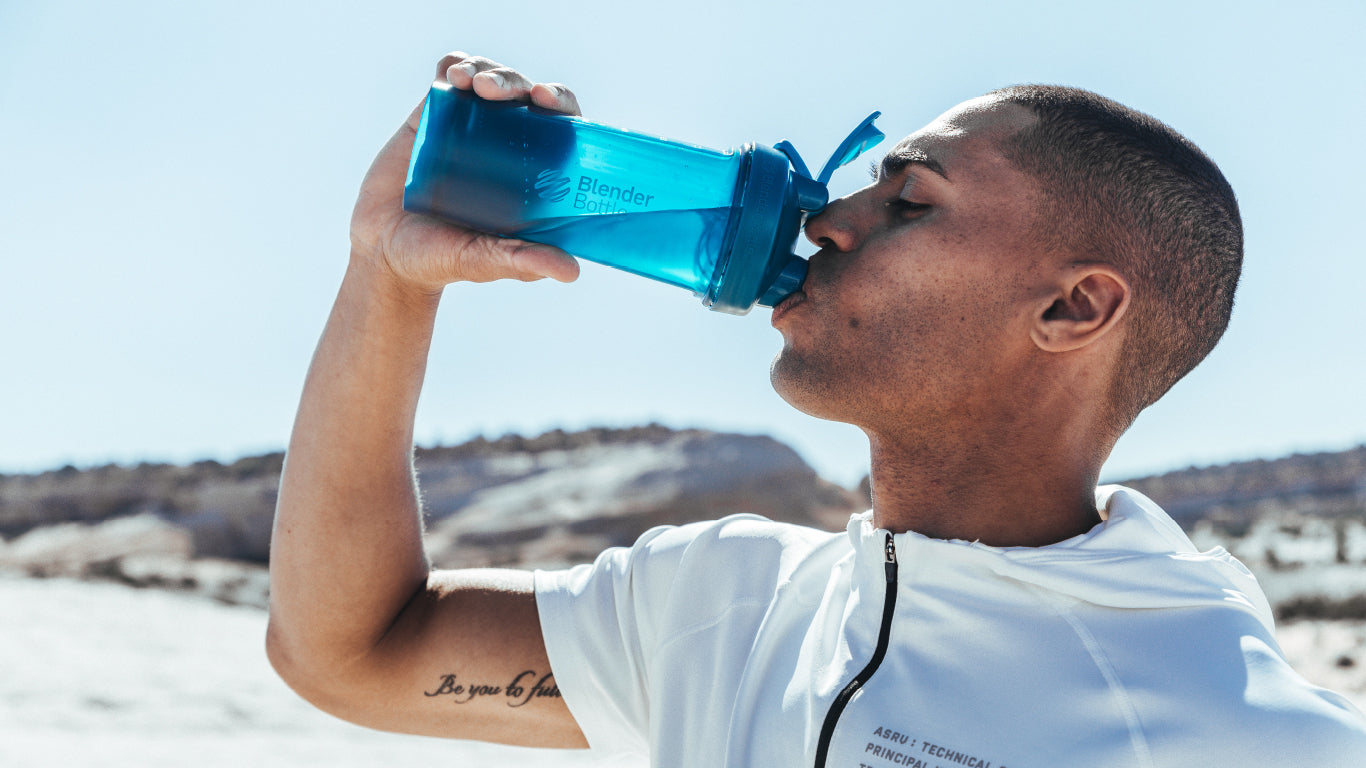 A man drinks from a blue BlenderBottle, showcasing the product's ergonomic design and vibrant color, with a focus on its practical use.