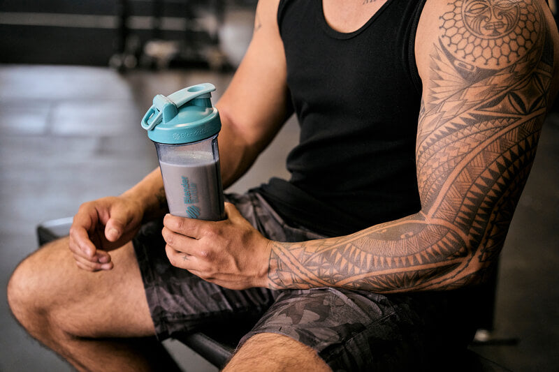 A tattooed man holding a shaker, emphasizing his muscular arms, suggesting an active lifestyle.