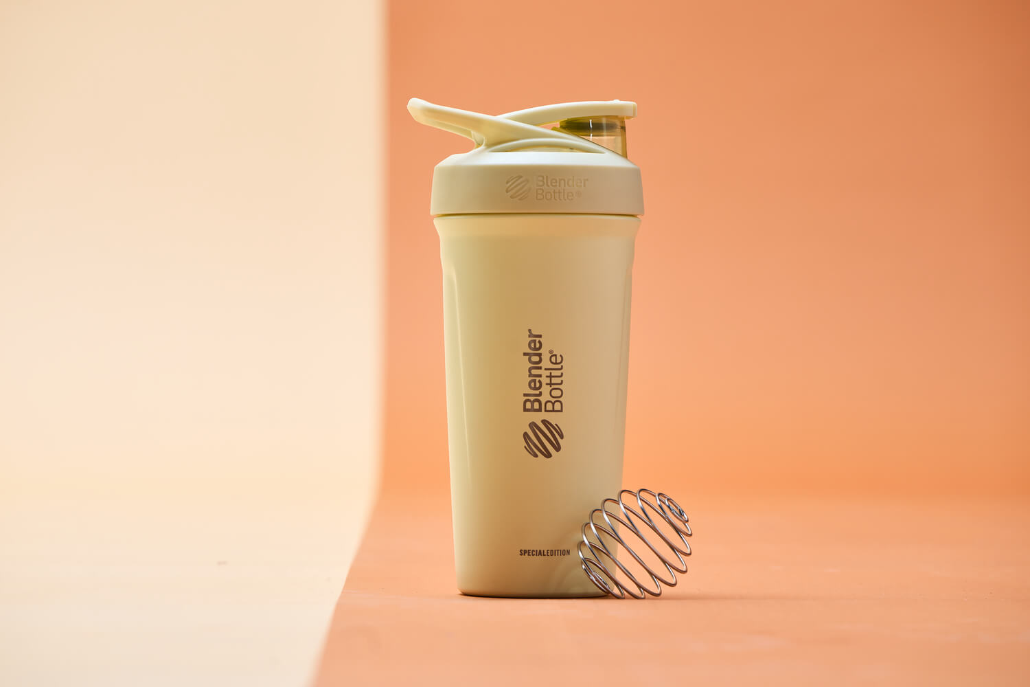 Cream stainless steel shaker with flip cap lid, featuring a metal BlenderBall wire whisk on an orange background.