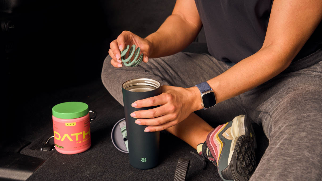 A person sitting on the floor holding a black BlenderBottle, and a green BlenderBall color wearing a black watch.