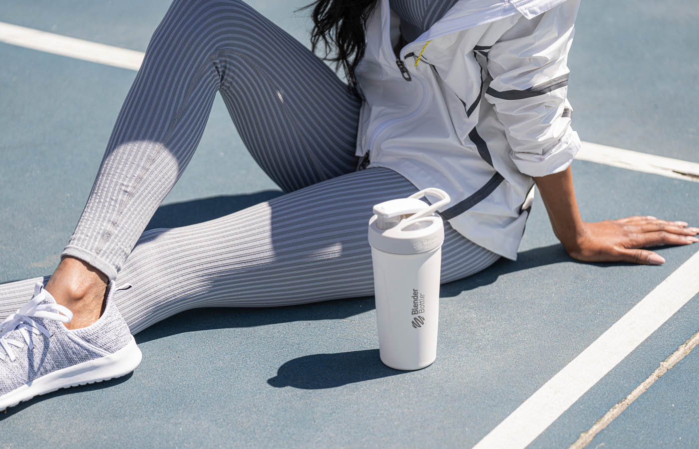 A woman sits on a tennis court holding a white water bottle, wearing casual clothing and sneakers.