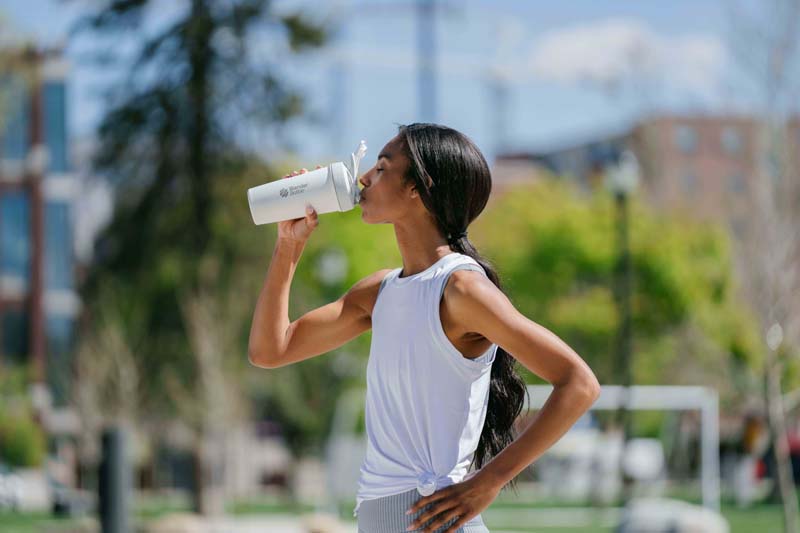 A woman drinks from a white BlenderBottle outdoors, surrounded by trees and grass, wearing casual clothing.
