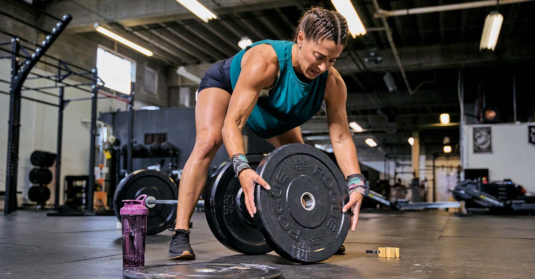 "A woman working out in a gym, with a purple BlenderBottle shaker bottle on the floor next to her.