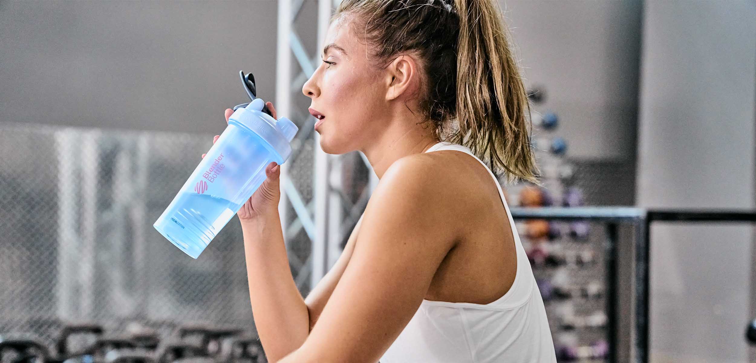 Woman in the gym drinking from a blue and pink BlenderBottle shaker cup, featuring a leak-proof design, a twist-on cap, and a BlenderBall wire whisk