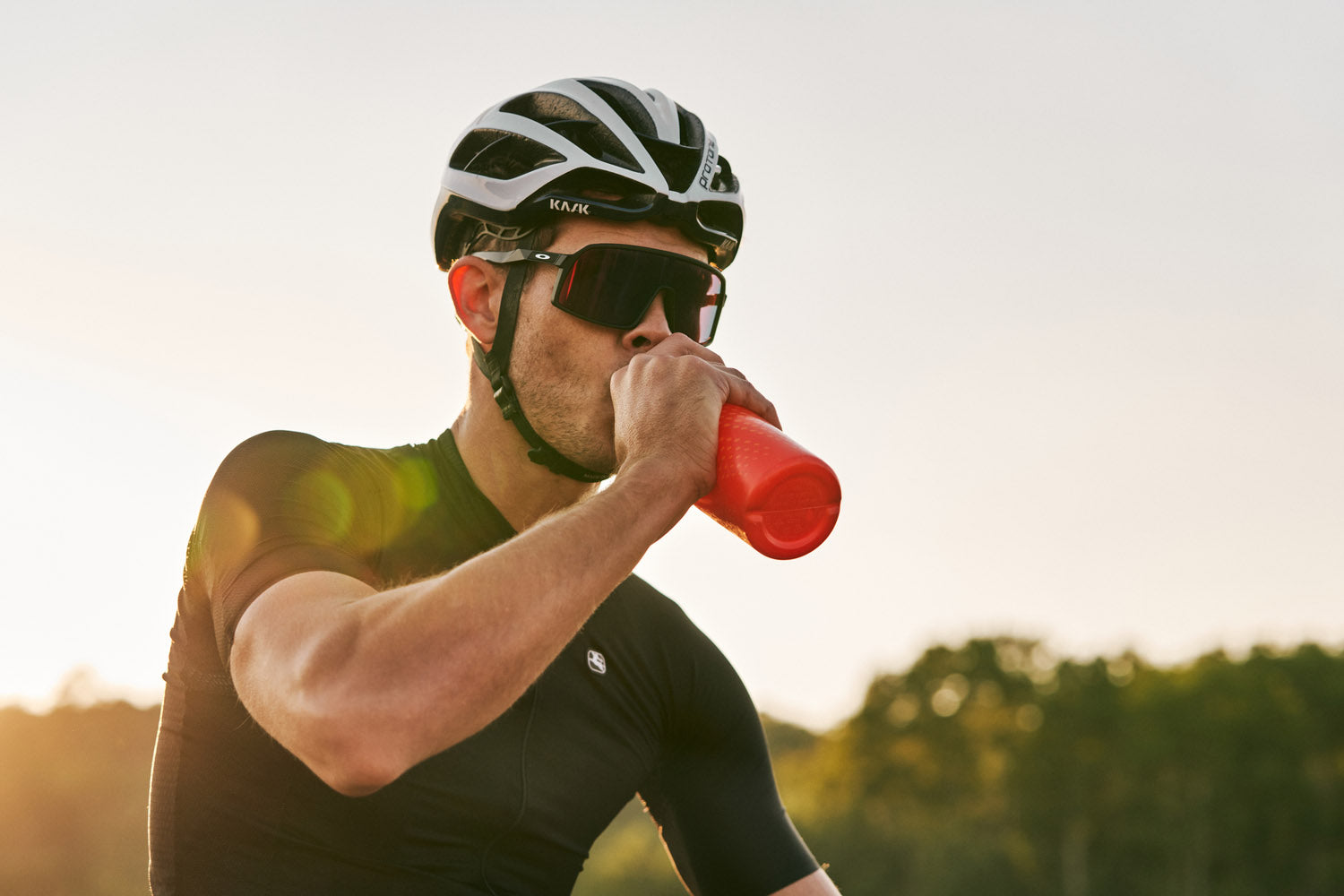 Man wearing a bicycle helmet and sunglasses drinks from a red bottle, suggesting a cycling activity or outdoor sports setting.