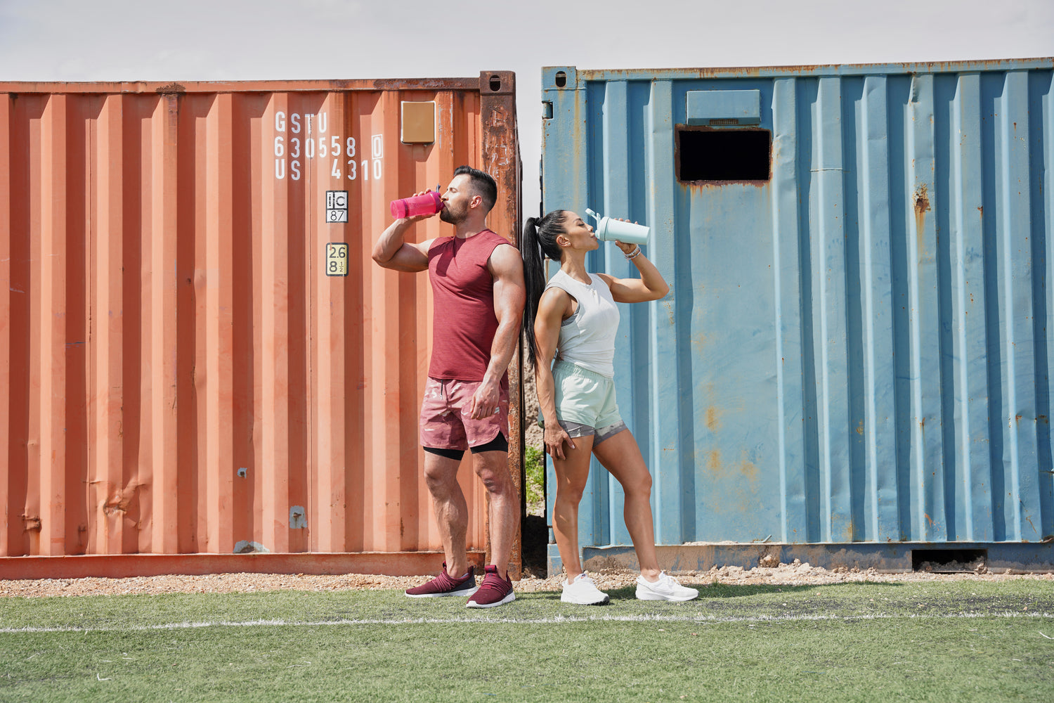 A man and a woman drinking from a BlenderBottles, standing on grass, wearing sneakers.
