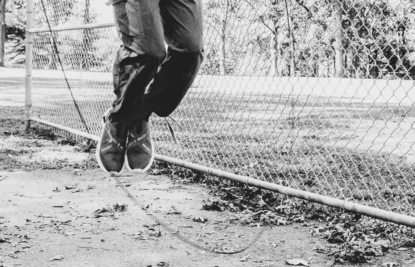 A person in black and white image jumps rope near a chain link fence, showcasing footwear during an outdoor activity.