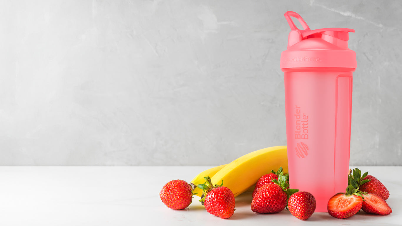 A pink BlenderBottle shaker surrounded by fresh strawberries and a banana, emphasizing a fruit-based shake concept.