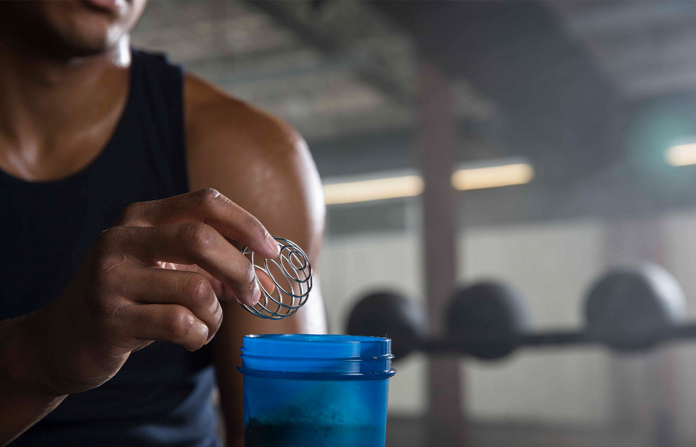 A person holds a BlenderBall wire whisk over a blue cup, suggesting preparation or mixing of a protein shake.