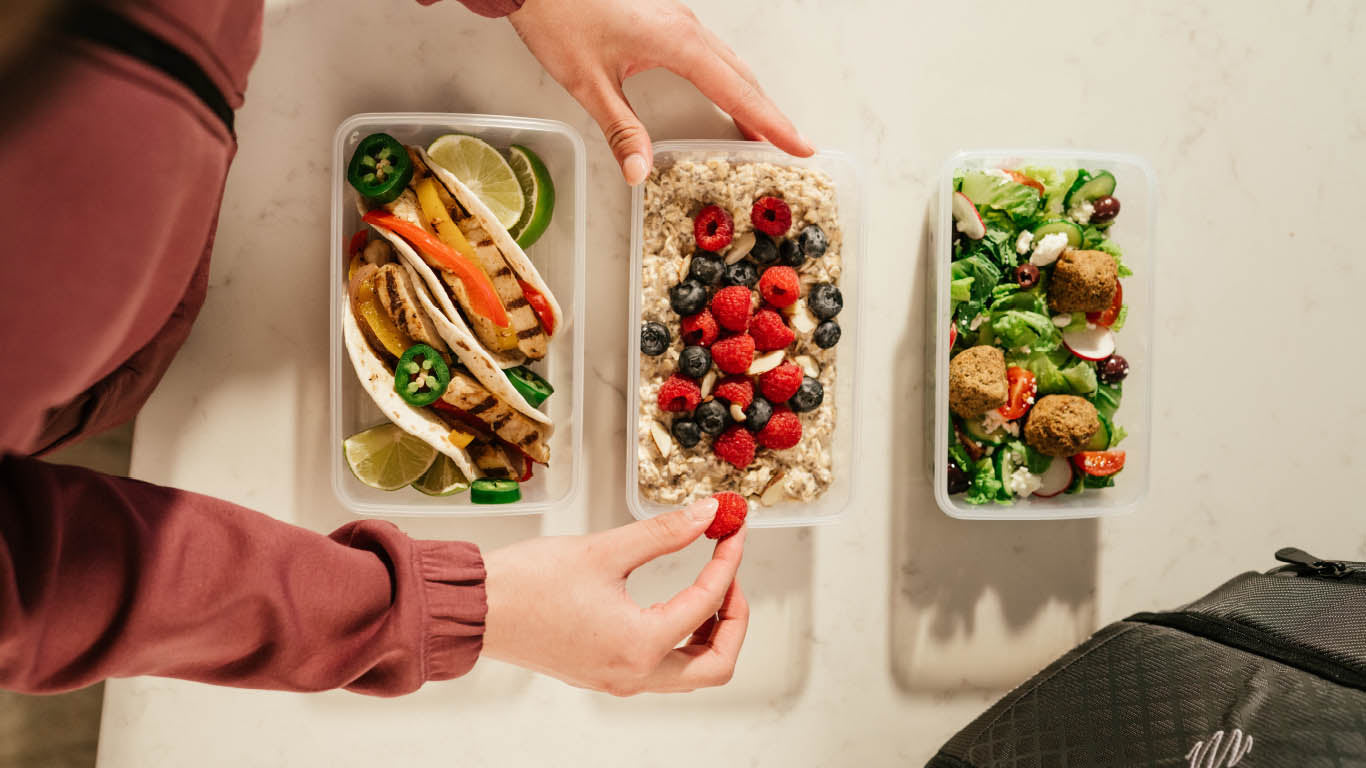 A person meal prepping containers of food featuring oatmeal with berries, a salad with meatballs and vegetables, and chicken tacos with vegetables.