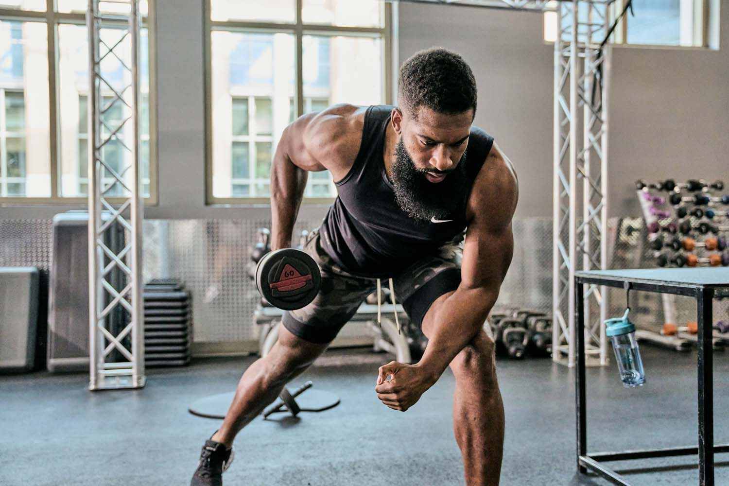 A man with a beard lifts weights in a gym, surrounded by exercise equipment and a water bottle on a nearby table.