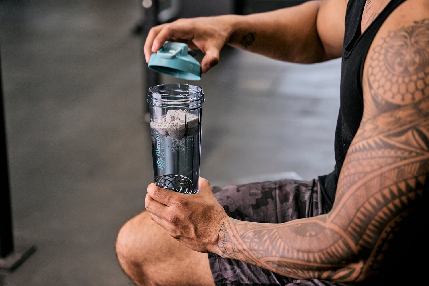 A man holding a blue-capped shaker bottle, with visible tattoos on his arm, in a gym setting. The shaker likely contains a protein mixture.