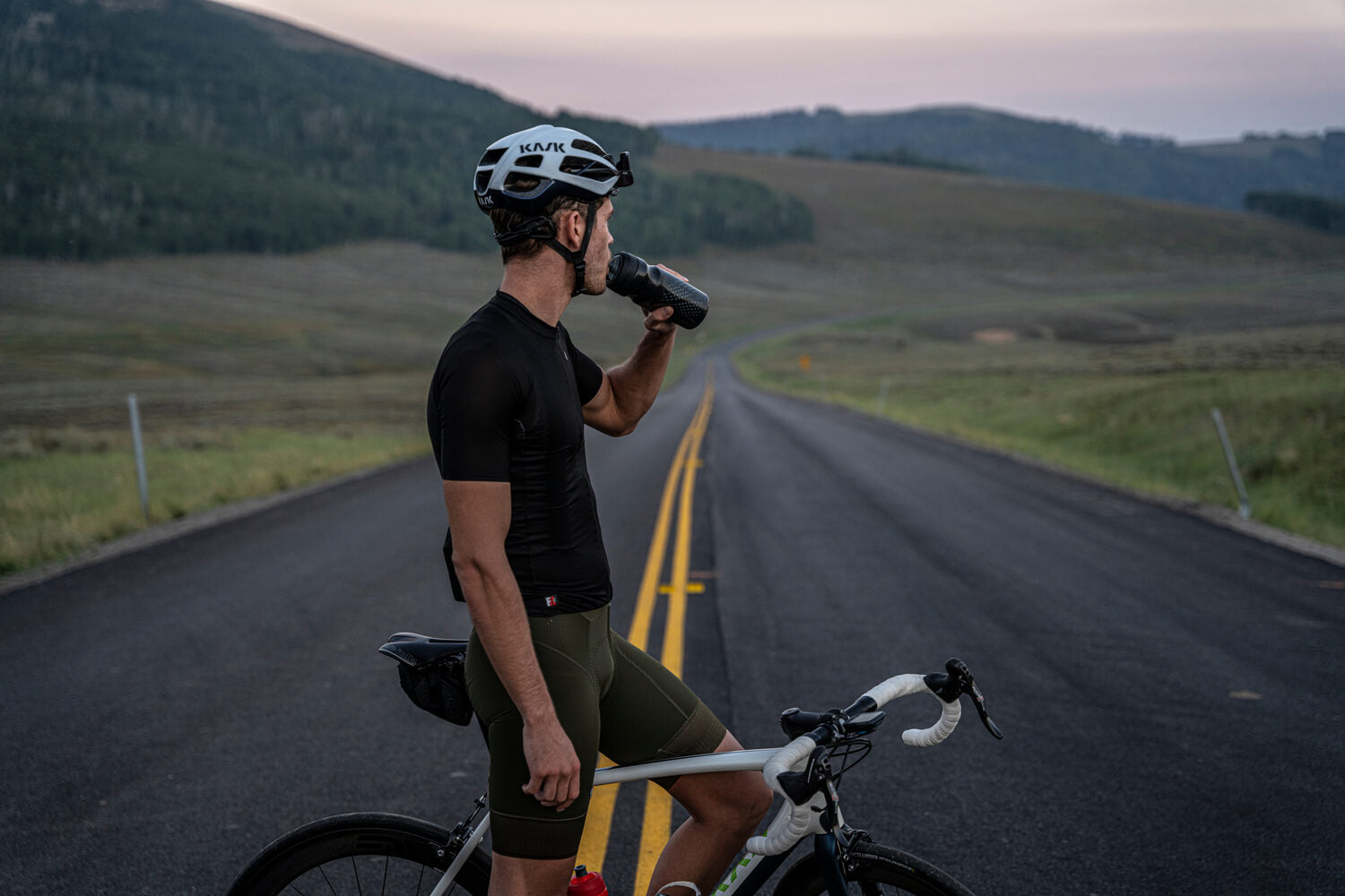 A man wearing a helmet drinks from a bottle while cycling on a road, with a mountain range visible in the background.