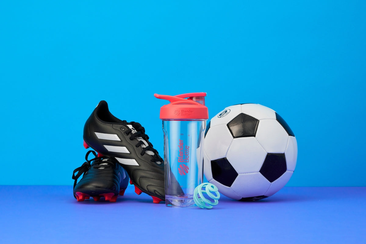 A close-up of a football, a BlenderBottle, and a pair of black cleats on a blue surface.
