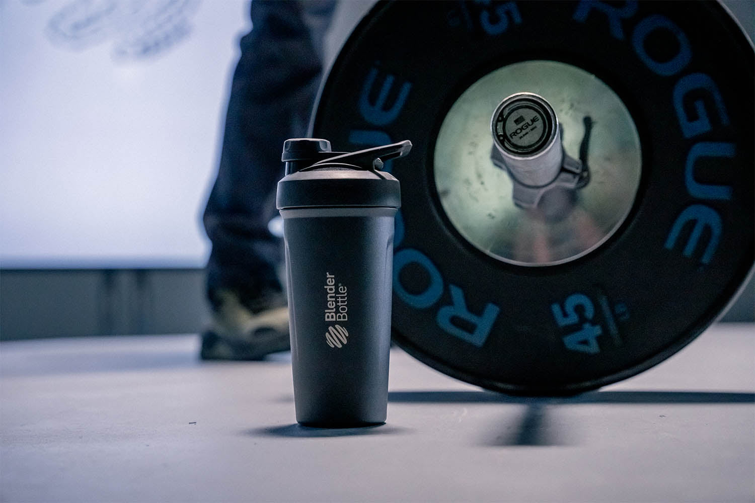 An insulated stainless steel shaker bottle with a twist lid on the floor next to a man working out