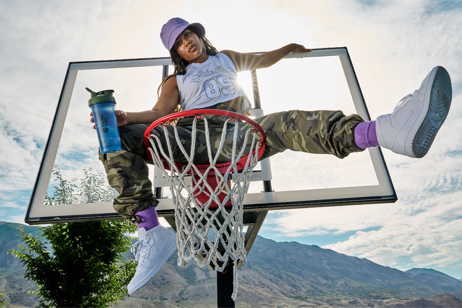 Woman holding a shaker cup sitting on a basketball rim.