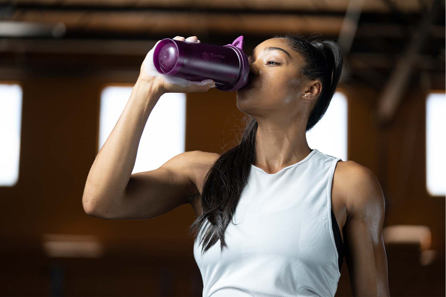 An image of a woman drinking out of a plum-colored BlenderBottle shaker cup in the gym. The woman is holding the cup with one hand and is seen taking a drink through the drinking spout. The cup has a leak-proof design, with a twist-on cap and a flip cap that snaps securely in place. 