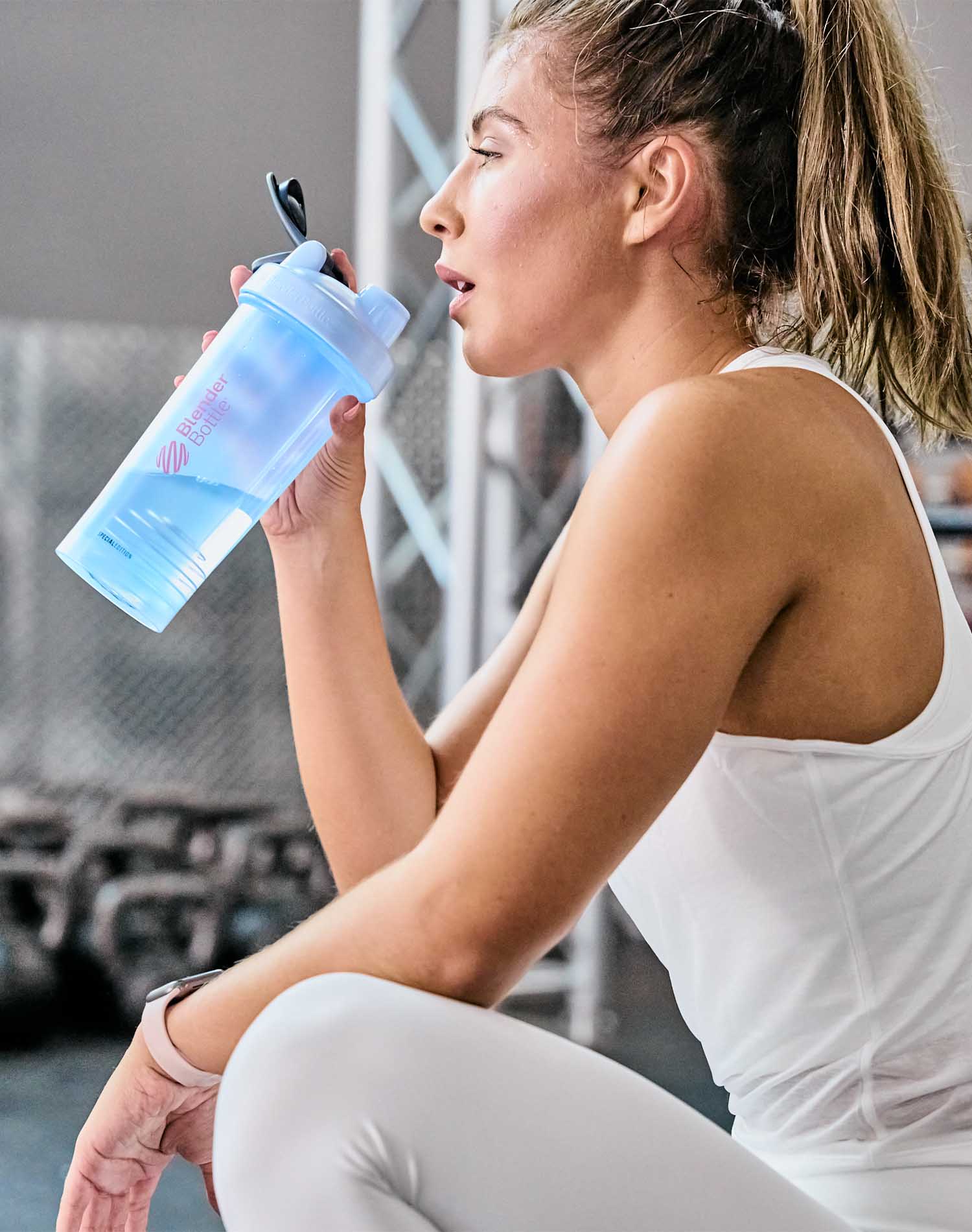 Woman in the gym drinking from a blue and pink BlenderBottle shaker cup, featuring a leak-proof design, a twist-on cap, and a BlenderBall wire whisk